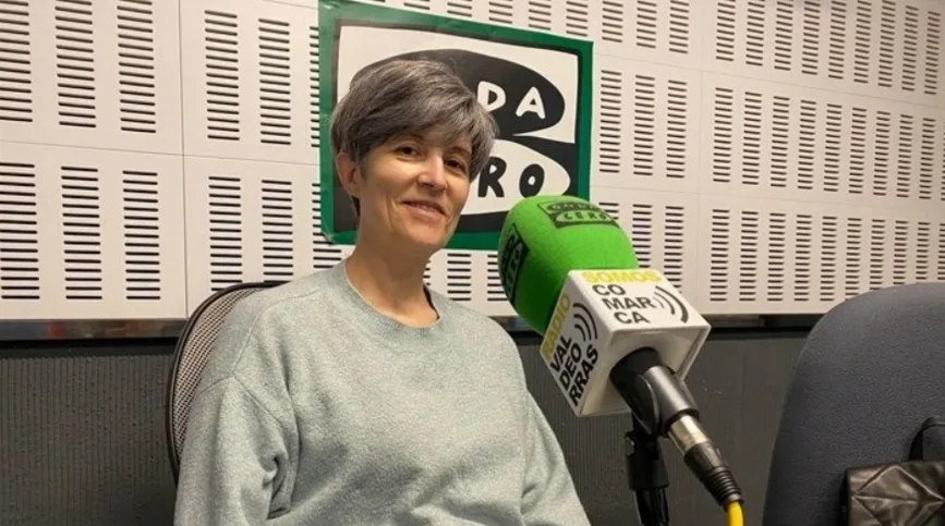 Ester, una mujer de cabello corto, sonríe sentada en un estudio de radio. Viste un jersey gris claro y se encuentra frente a un micrófono profesional con una funda de espuma verde de la emisora Onda Cero. El soporte del micrófono muestra el logotipo de Somos Comarca. Al fondo se aprecia una pared con paneles de insonorización blancos y un cartel corporativo.